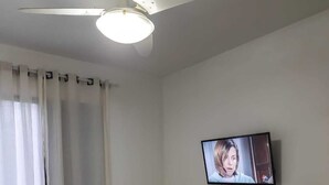 A bedroom with a bed, a television on the wall, a ceiling fan, and a window with curtains.