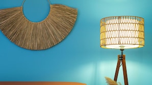 Un lit recouvert d’une couverture marron et agrémenté d’oreillers à motifs, une décoration murale tissée et une lampe de sol avec un pied blanc et un abat-jour en tissu tressé.