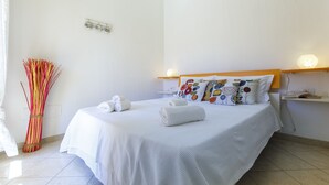Een netjes opgemaakt bed met witte lakens en decoratieve kussens, een nachtkastje met een lamp, en een wandlamp.