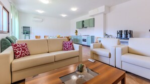 A modern living room with a beige sofa, a wooden coffee table, and a glass-top table with a cactus. There is a kitchen in the background with green cabinets and a stove. - Countryside, nature e private swimming pool (Carbonia)