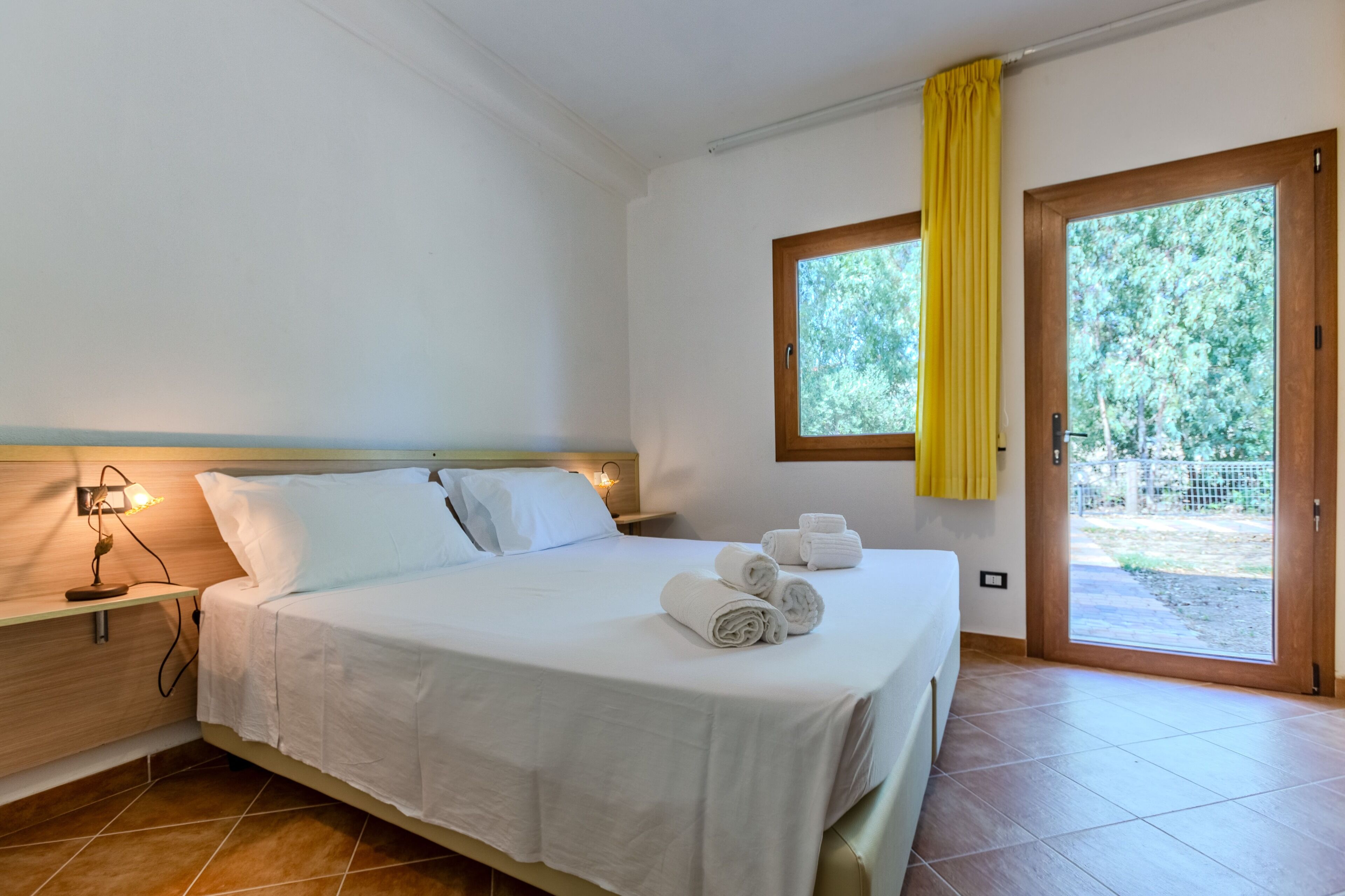 A clean, modern bedroom with a bed, bedside lamp, and a view of greenery through a window.