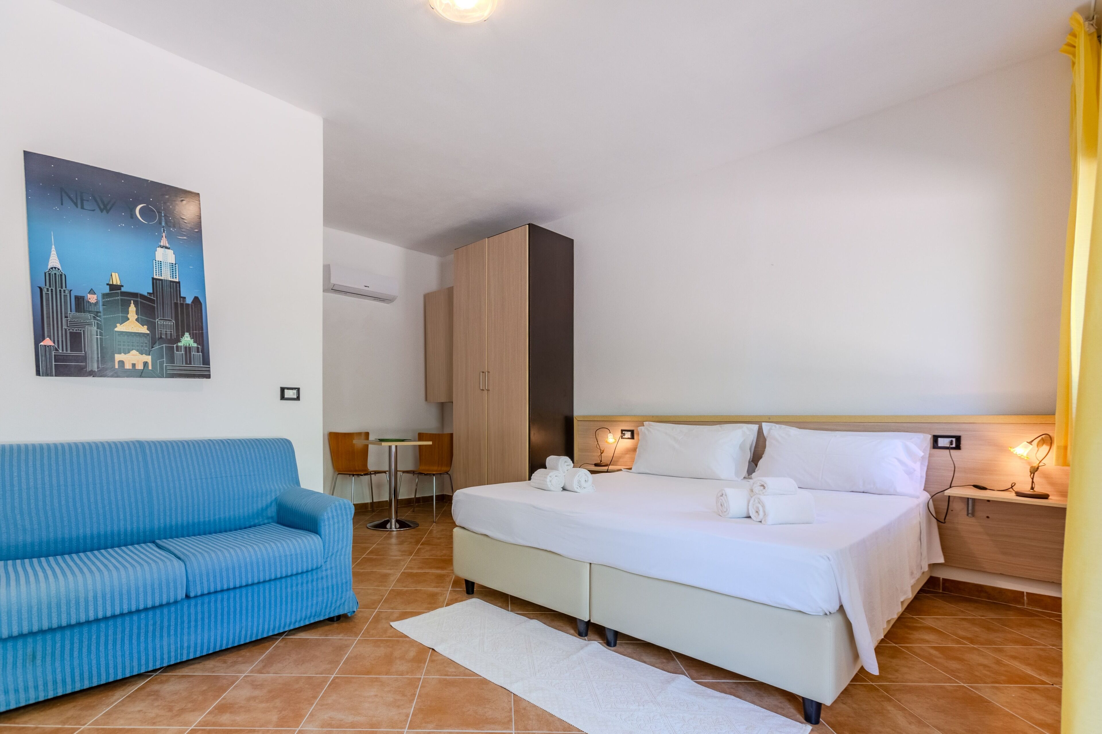 Habitación de hotel con cama, sofá azul, escritorio y cuadro de un paisaje urbano en la pared.