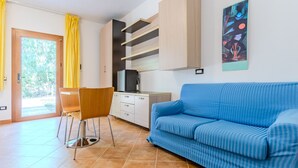 A modern living room with a blue sofa, a dining table with chairs, a kitchen area with a refrigerator, and a sliding glass door leading to a balcony. - Countryside house - 2 (Carbonia)