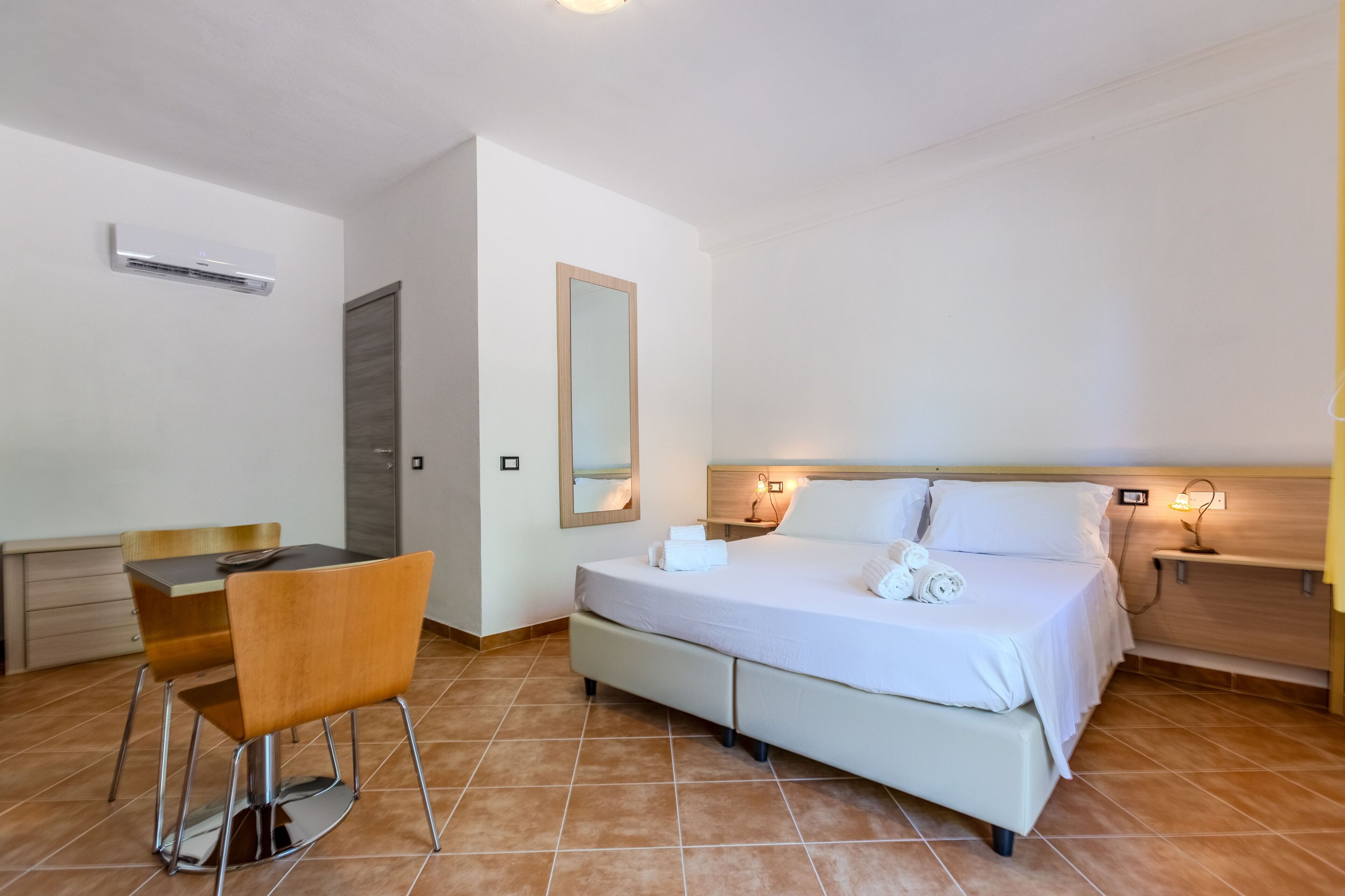 Quarto de hotel com cama, mesa de cabeceira, cadeira e pequena escrivaninha.