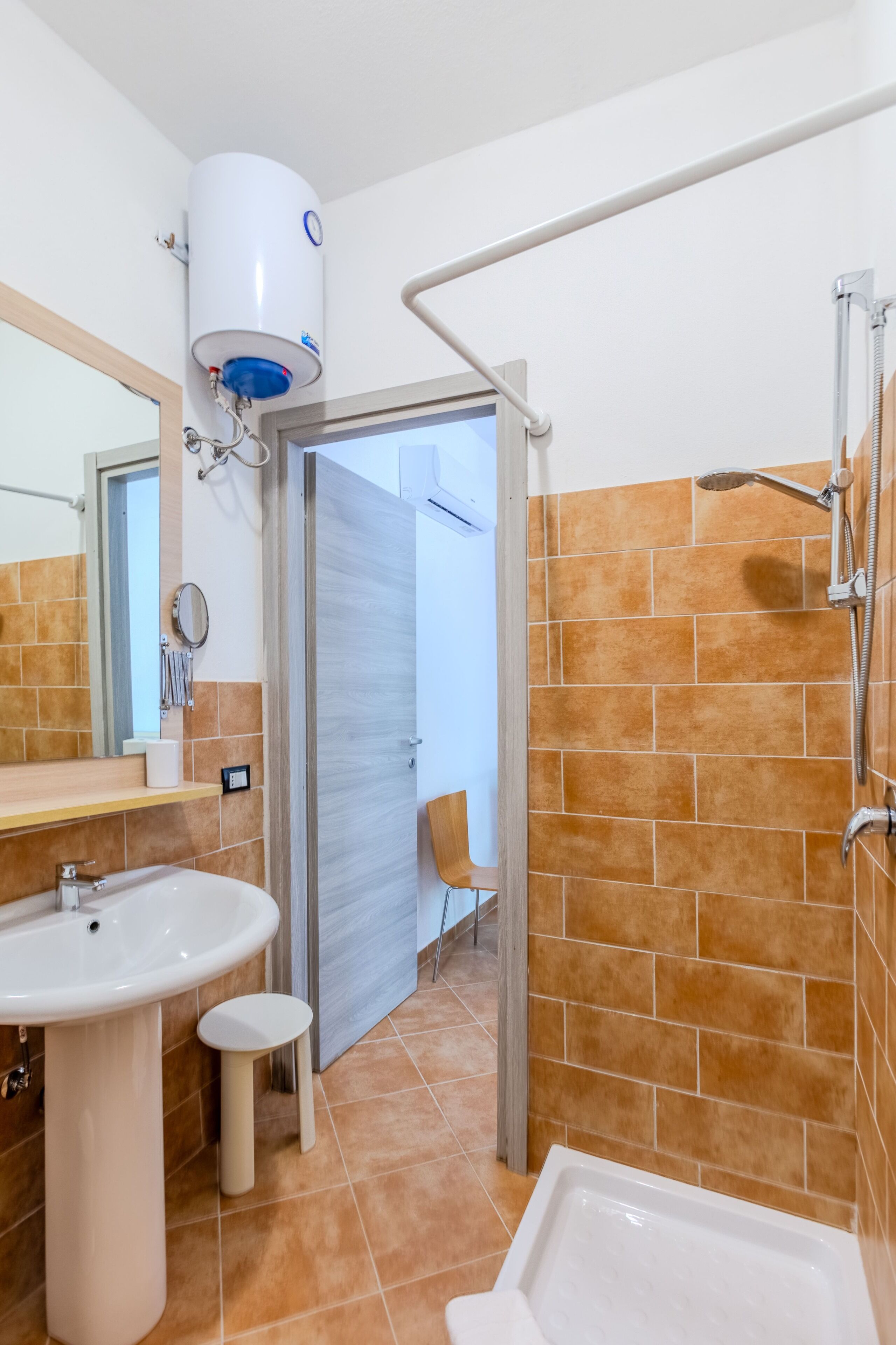 A bathroom with a white sink, a small stool, a shower with a handheld showerhead, and a white toilet.
