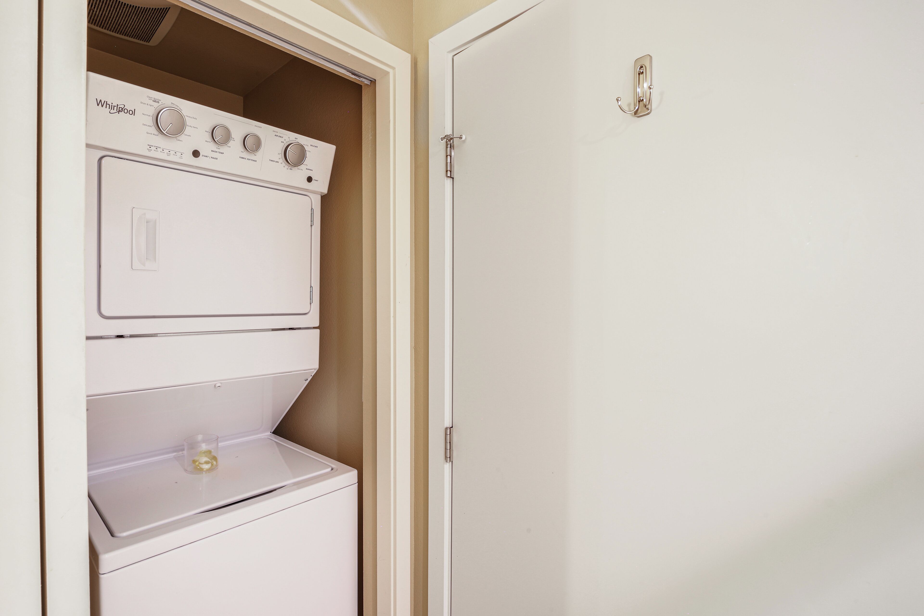A laundry room with a stacked washer and dryer, a door with a light switch, and a small shelf with a glass container.