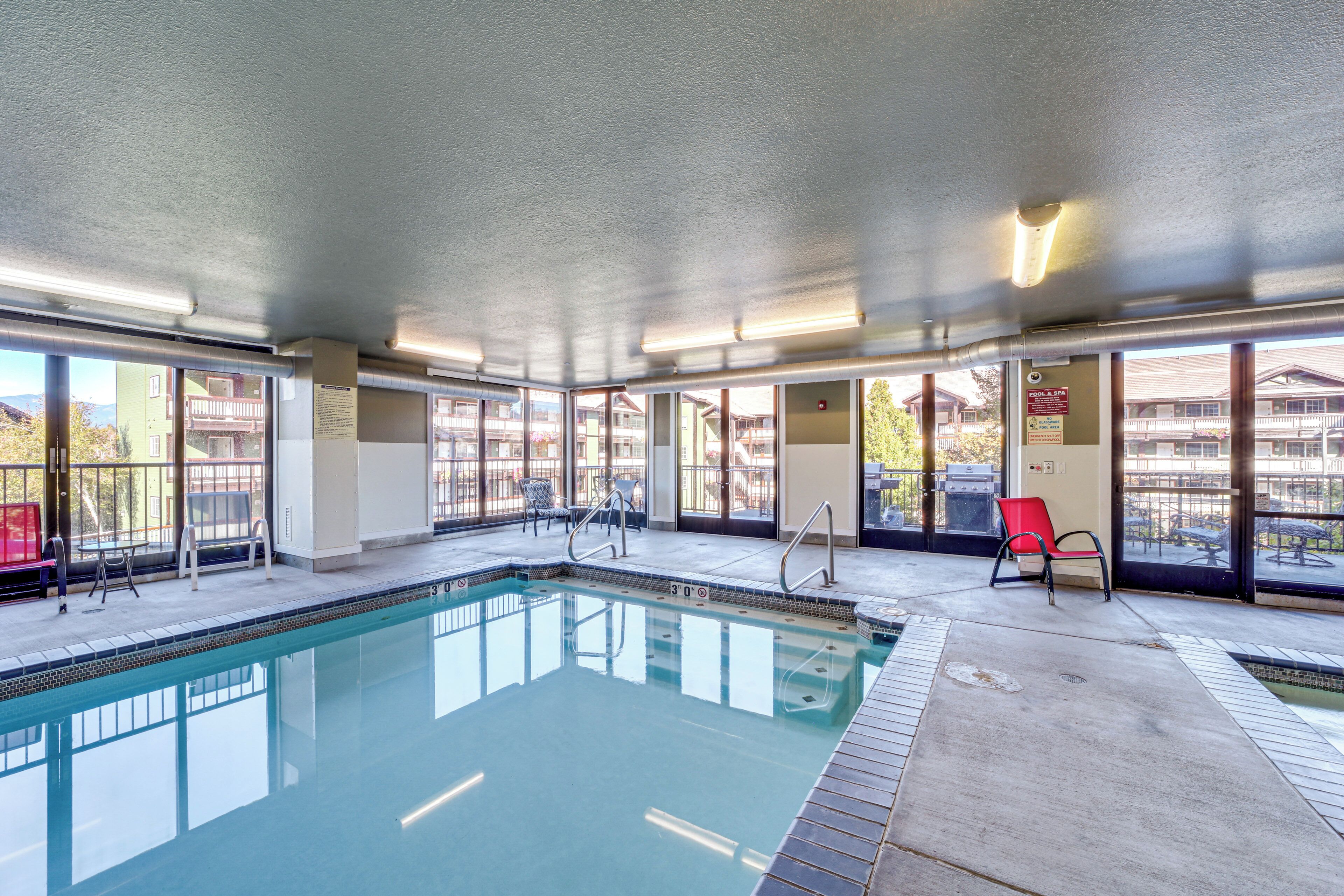 An indoor swimming pool with clear glass walls, a tiled floor, and a few chairs around the pool area.