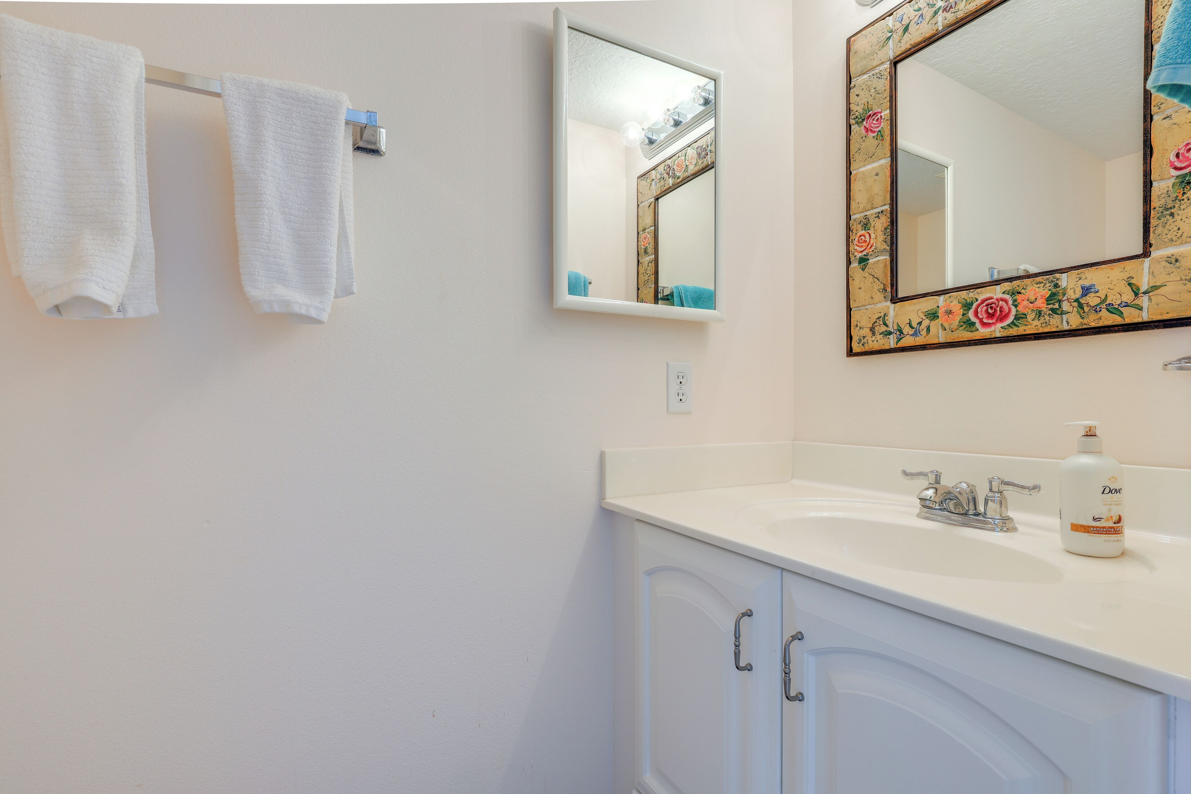 A bathroom with a white sink, a mirror with a floral frame, and two white towels hanging on a rack.
