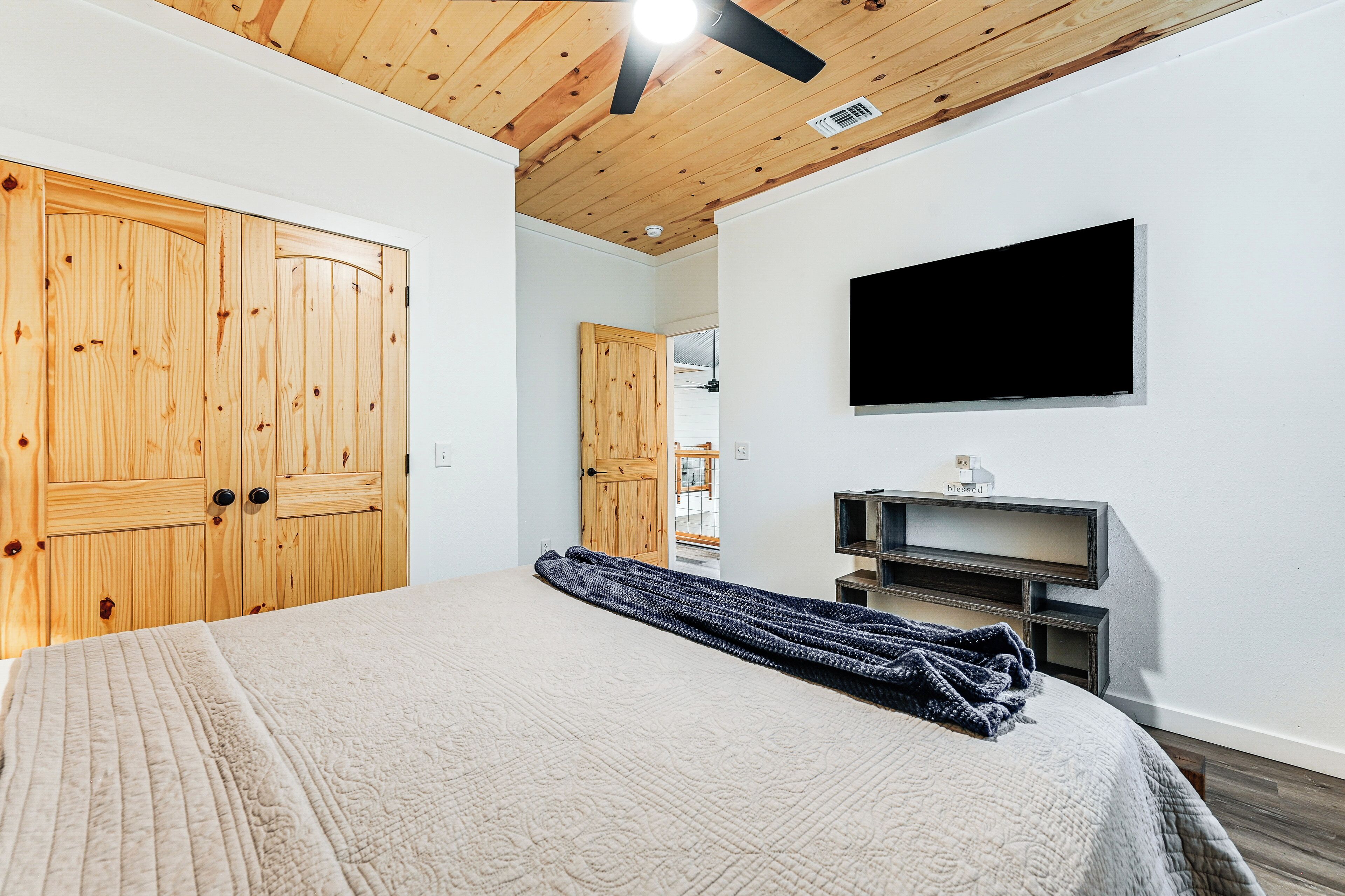 A bedroom with a large bed, wooden wardrobe, a mounted TV, and a ceiling fan.