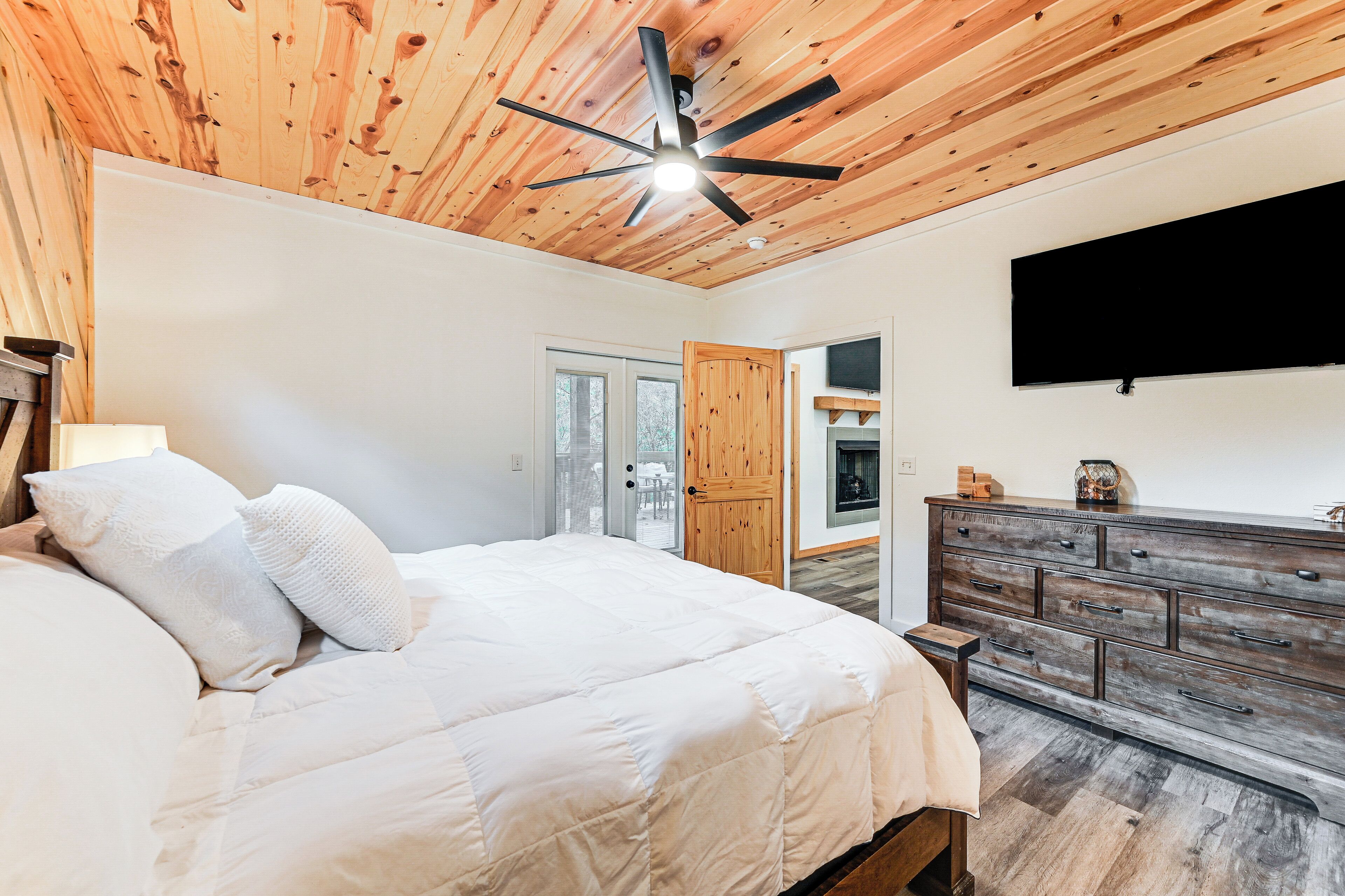 A bedroom with a wooden ceiling, a bed with white bedding, a dresser, and a TV mounted on the wall.