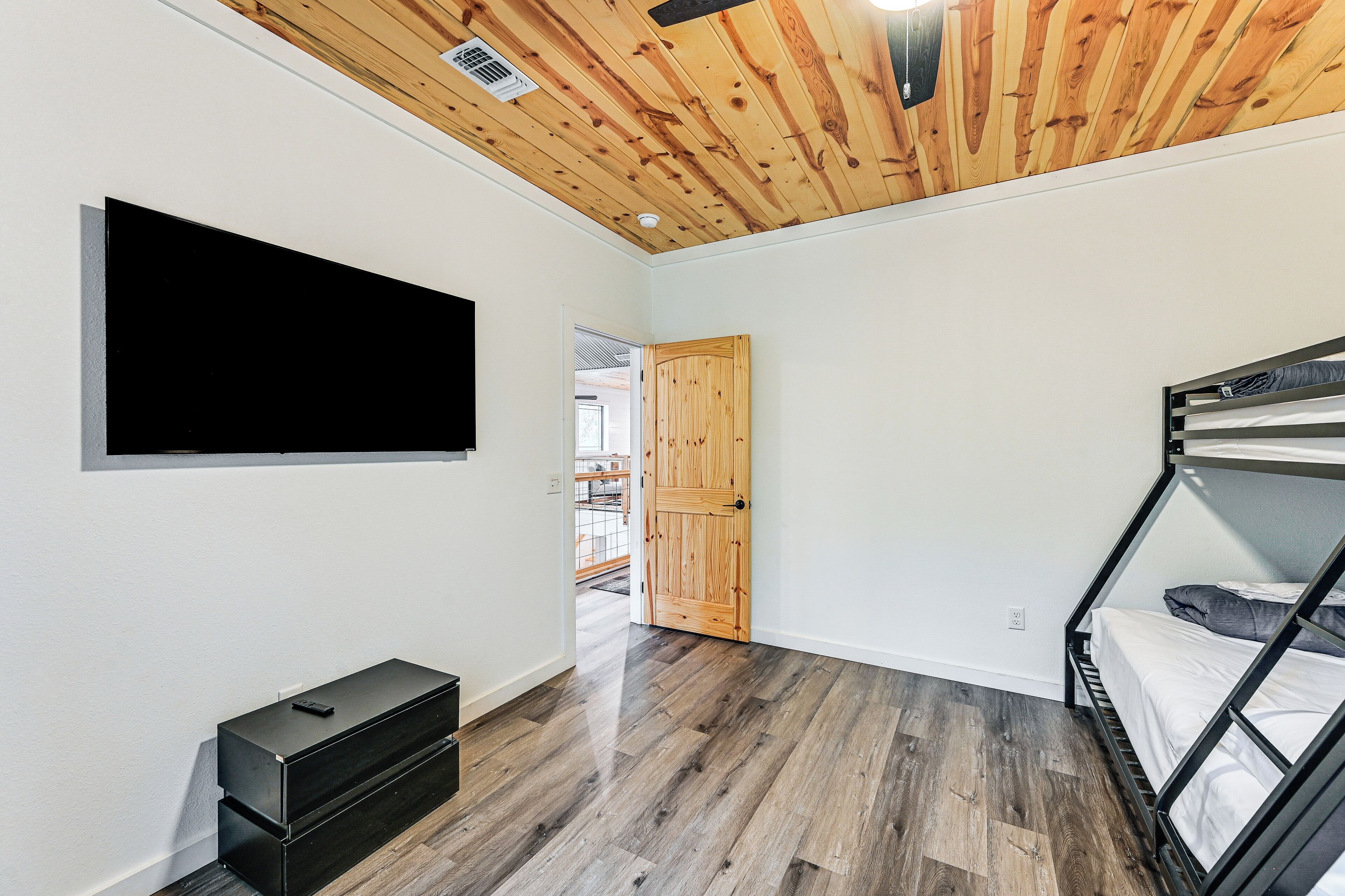 A room with a wooden ceiling, a bed with a metal frame, a TV mounted on the wall, and a small black cabinet.