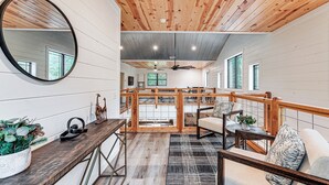 Ein geräumiges Zimmer mit Holzbalkendecke, einem großen Spiegel, einer hölzernen Bank, einem kleinen Tisch mit Pflanze und Blick zur Küche durch eine Glastür.