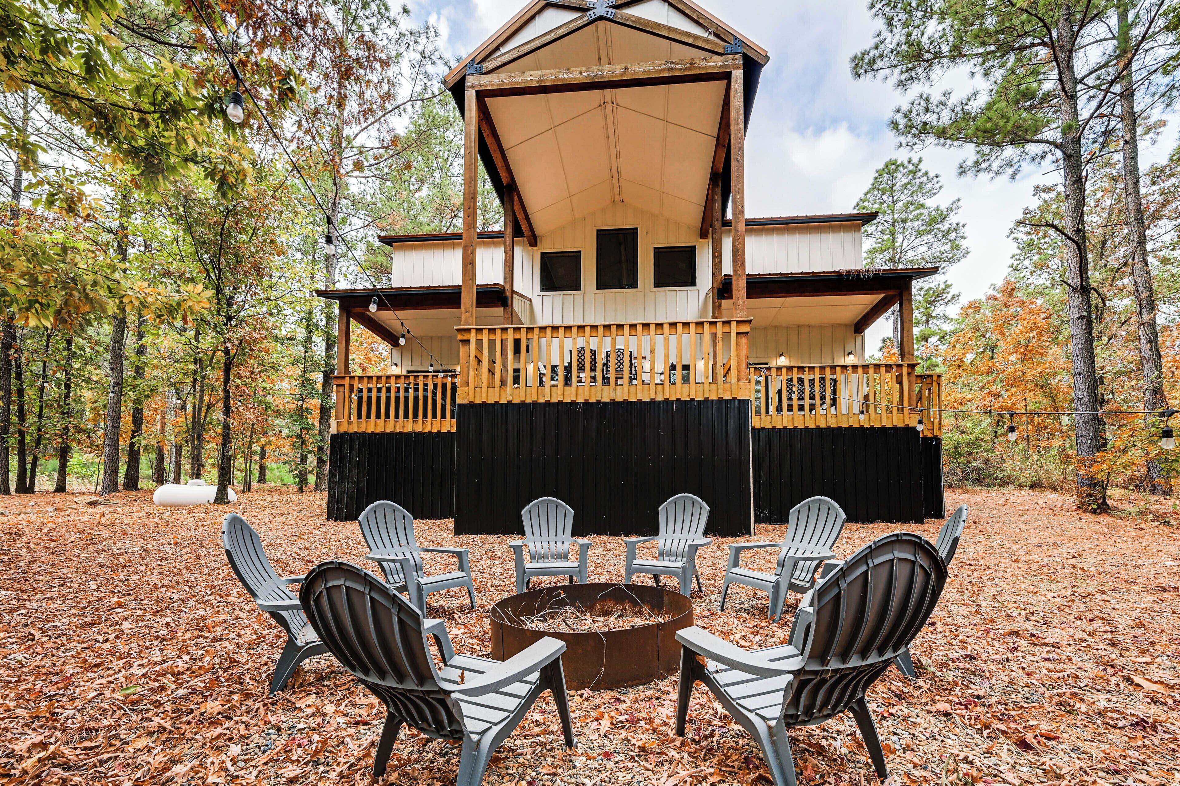 A cabin with a porch, surrounded by chairs and a fire pit, set in a wooded area.