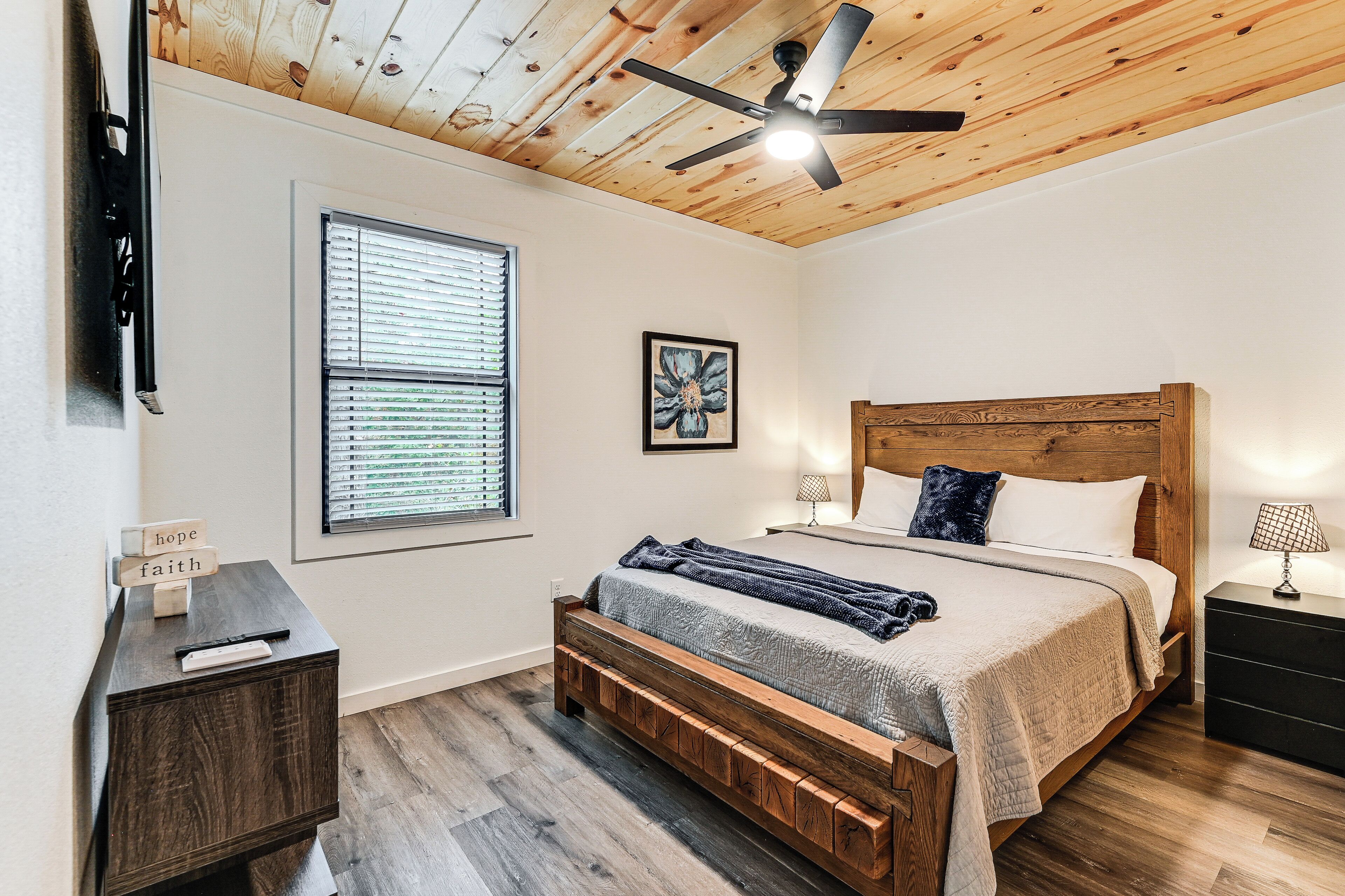 A bedroom with a wooden ceiling, a bed with a headboard, a bedside table, a nightstand, a window with blinds, and a ceiling fan.