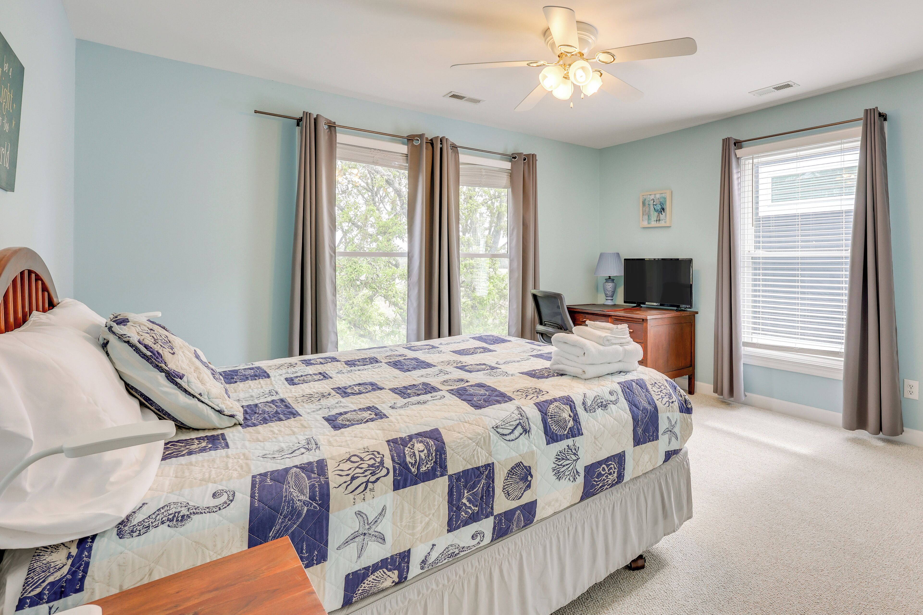 A bedroom with a bed, a desk with a computer, and two windows with curtains.