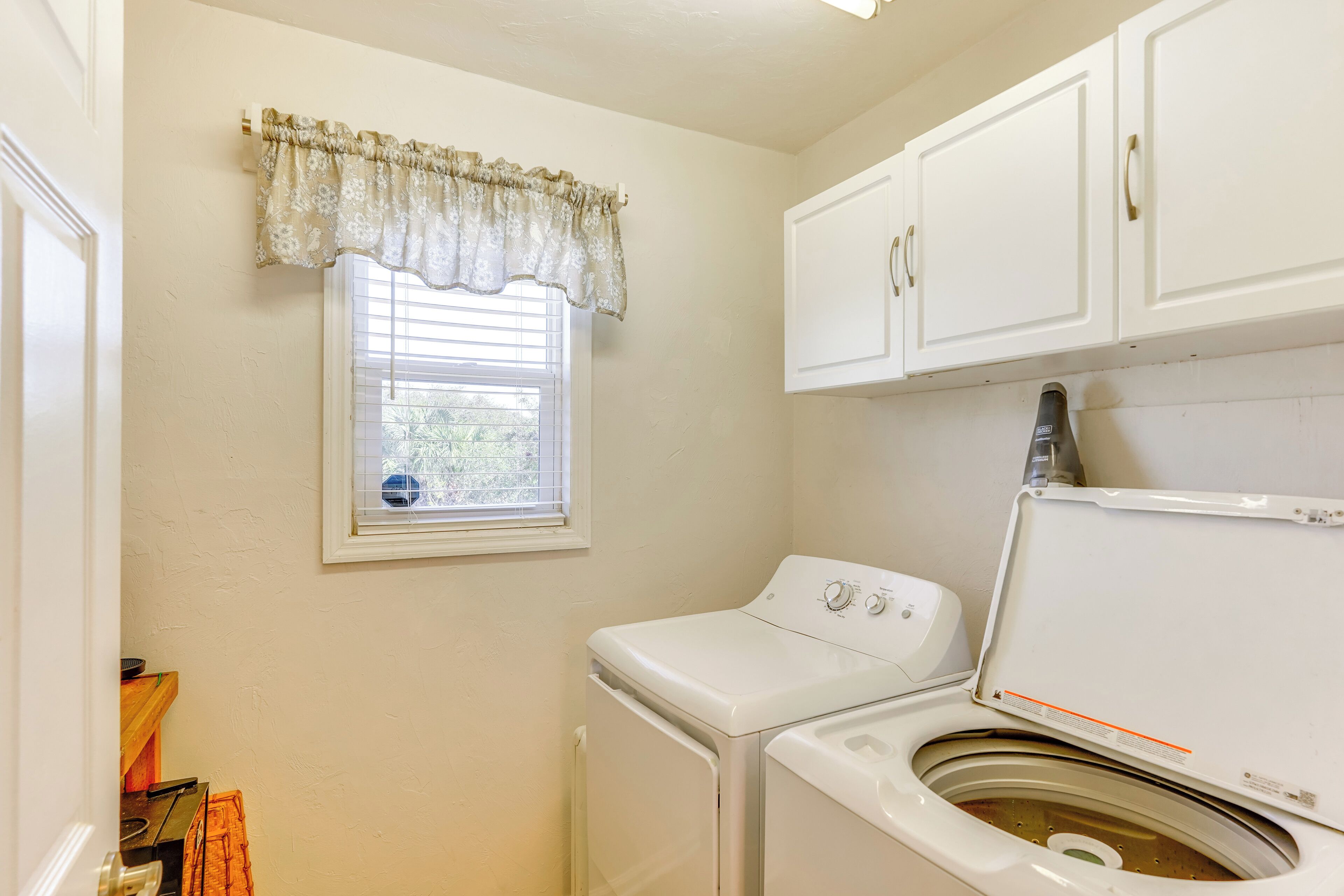 A laundry room with a front-loading washer and dryer, a window with a valance, and white cabinets above.