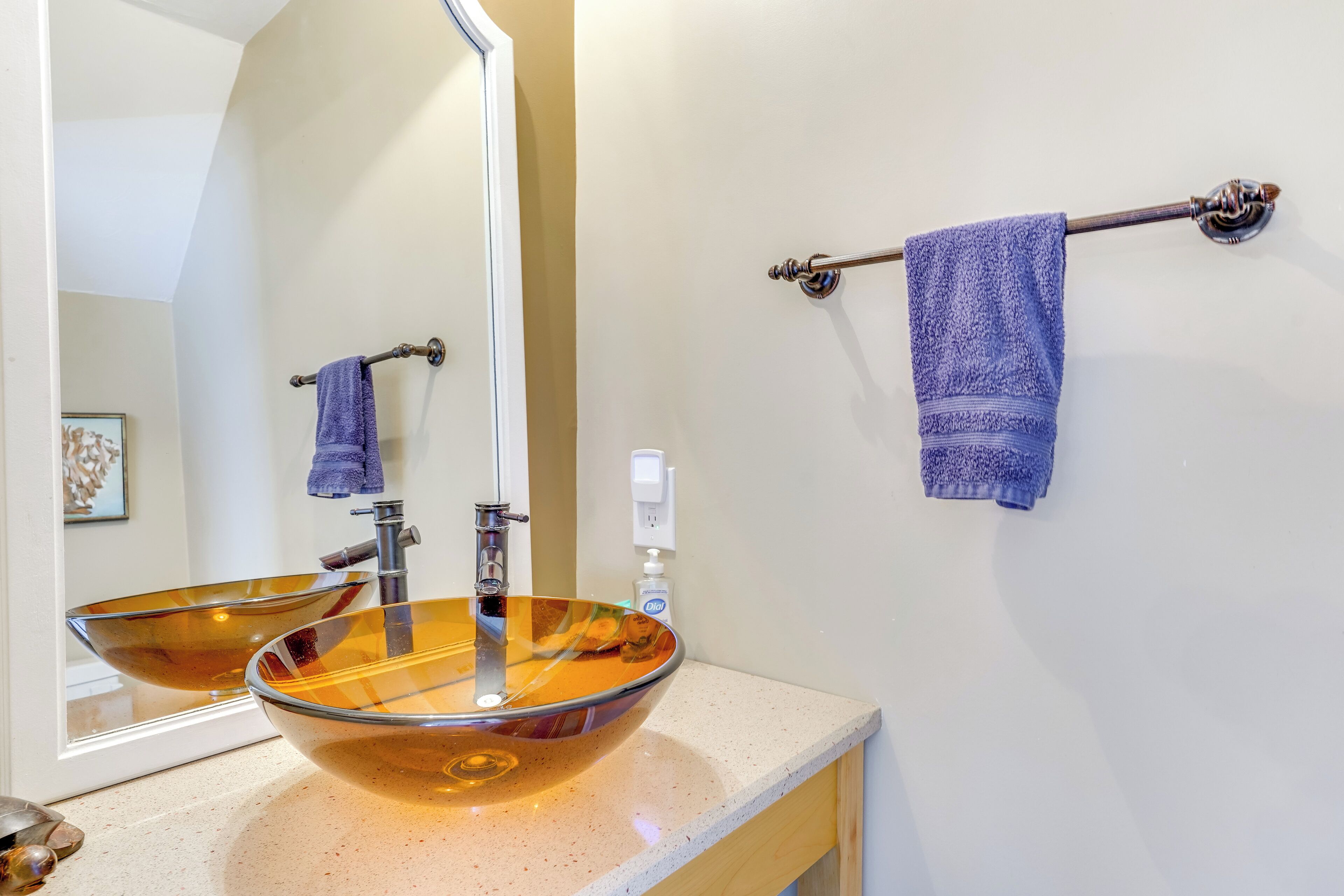 Une salle de bain avec un grand lavabo en forme de bol en verre, un miroir, un porte-serviettes avec une serviette bleue et un distributeur de savon.