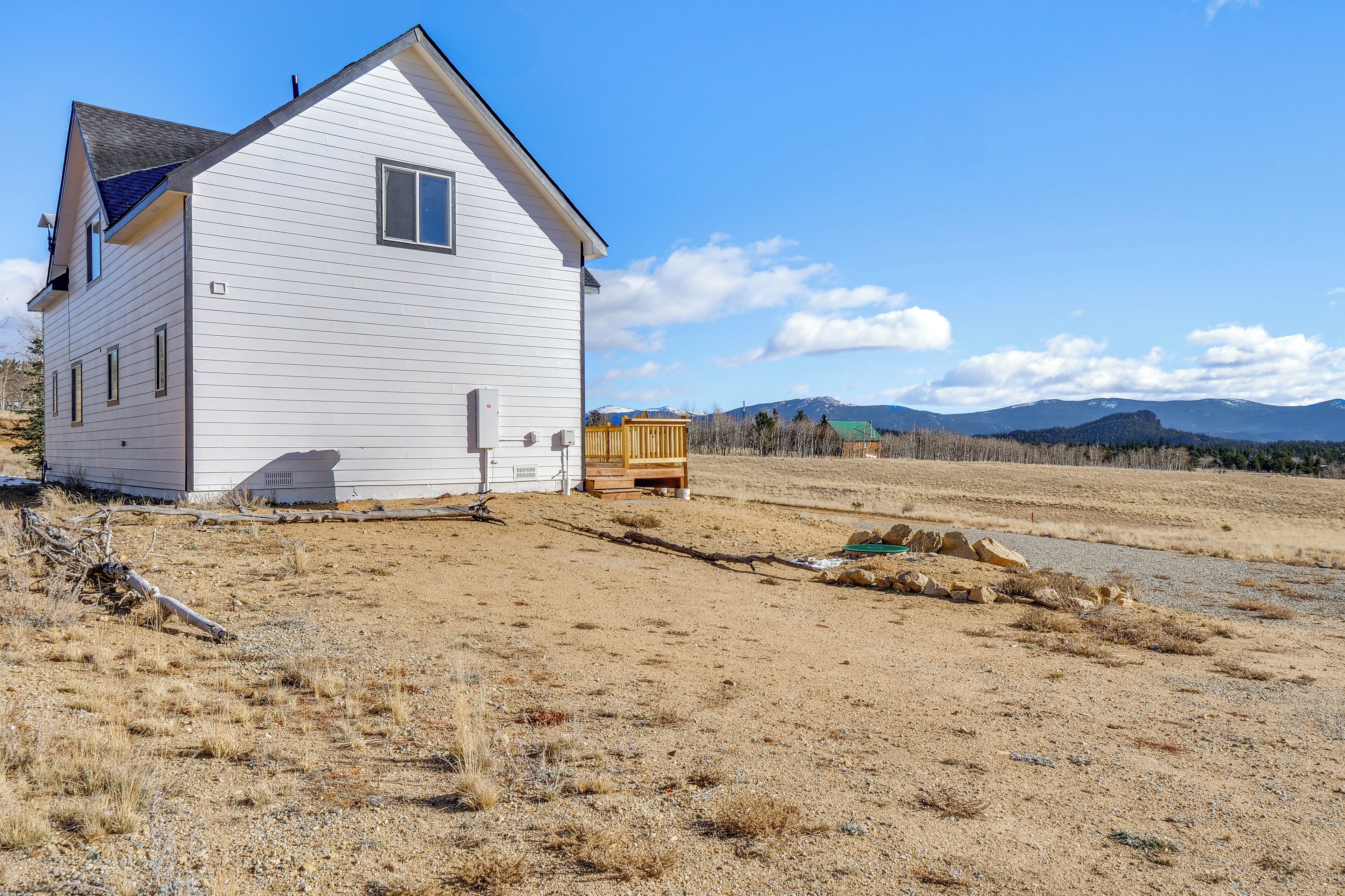 A white house with a sloped roof, a wooden deck, and a dry, grassy field with mountains in the background.