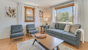 A living room with a grey sofa, two armchairs, a wooden coffee table, and a large window with curtains.