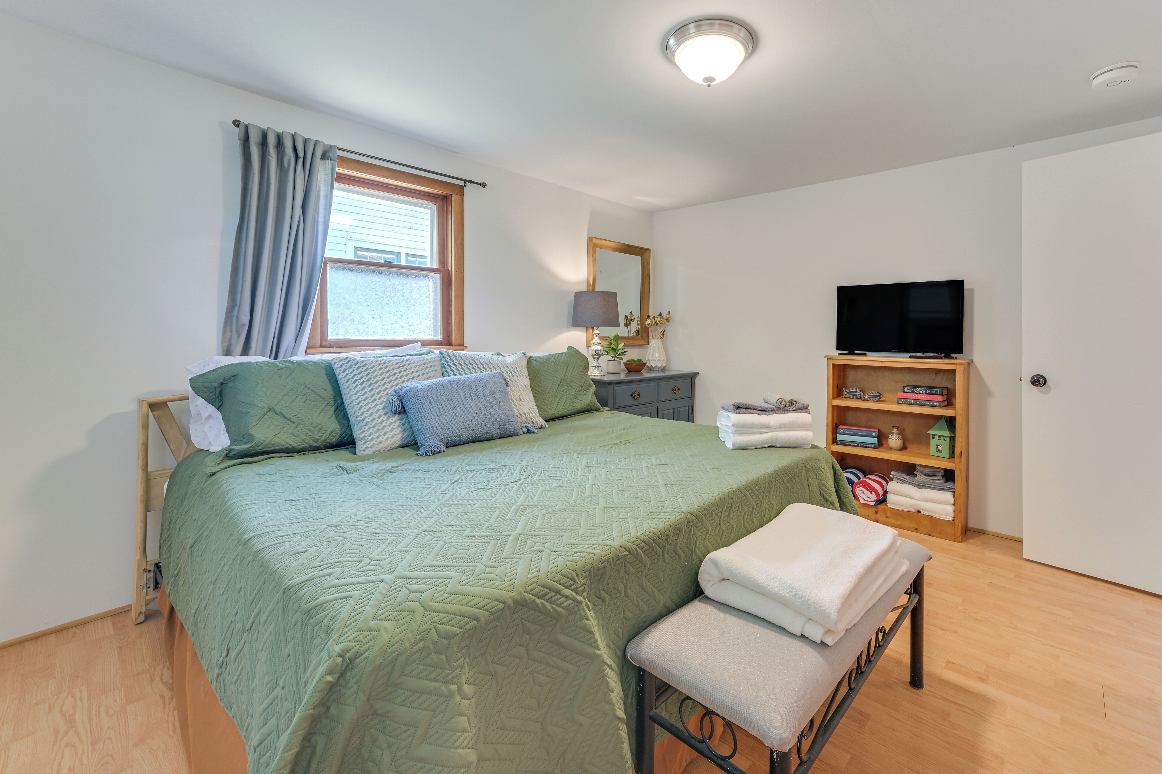 Une chambre à coucher avec un lit, un banc, une télévision, une bibliothèque et une fenêtre avec des rideaux.