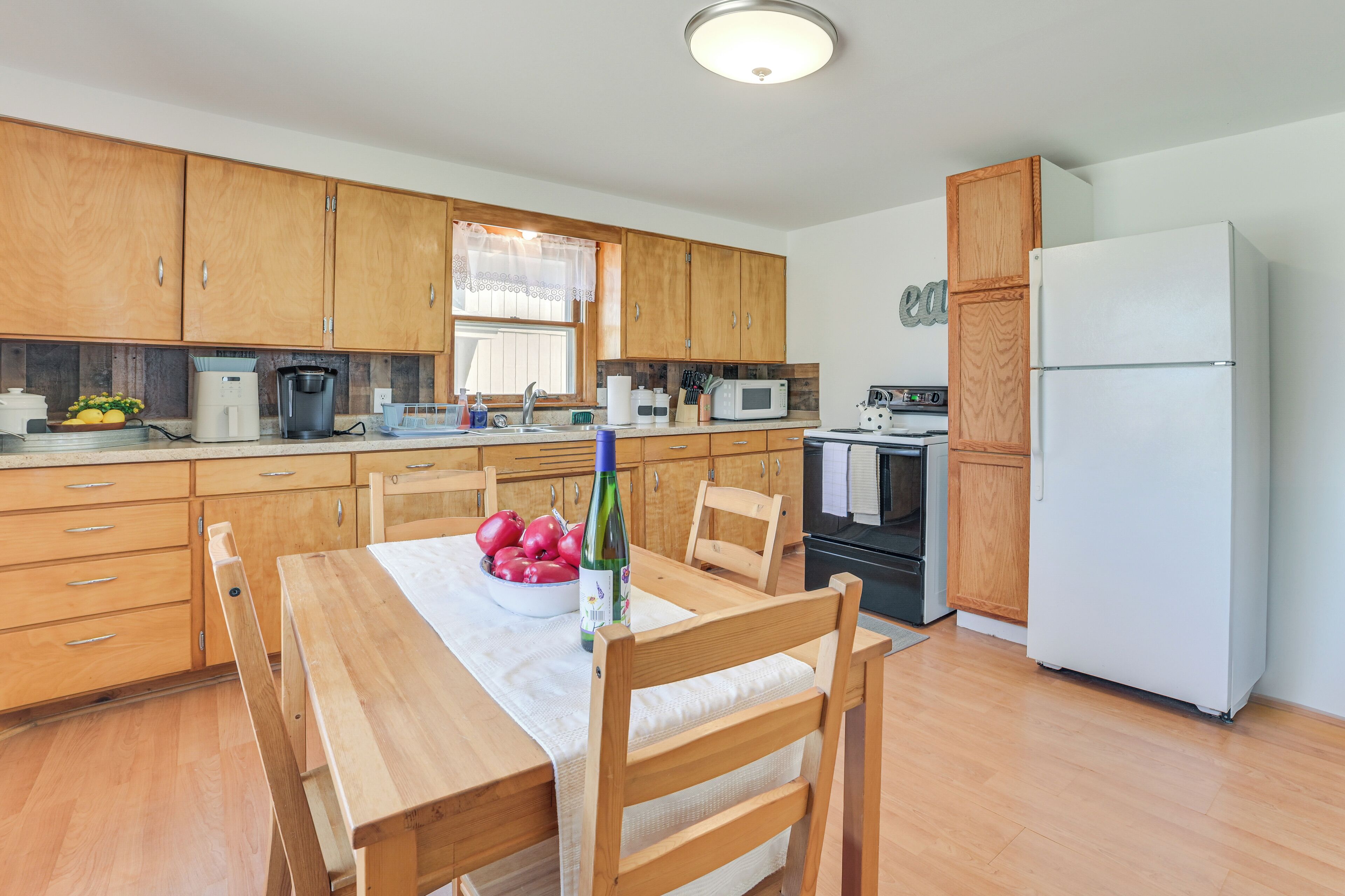 A modern kitchen with wooden cabinets, a white refrigerator, a dining table with chairs, and a bowl of fruit.