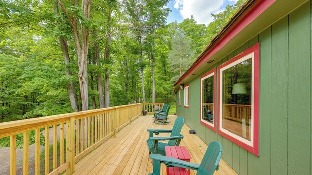 Una terraza de madera con sillas Adirondack verdes y una mesa roja, con vistas a una zona boscosa.