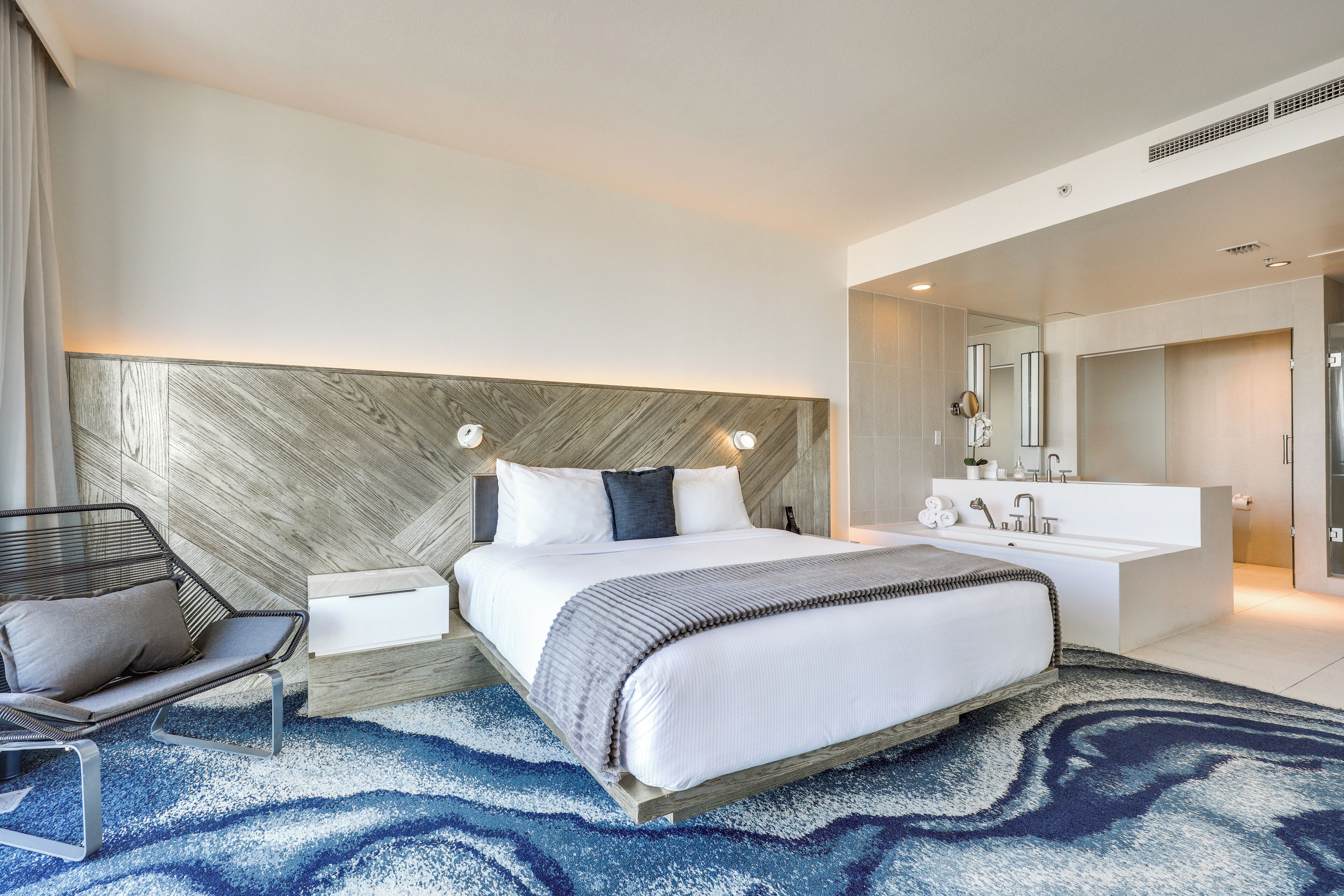 Quarto de hotel moderno com uma cama grande, mesa de cabeceira, uma cadeira, toucador embutido com espelho e um tapete azul estampado.