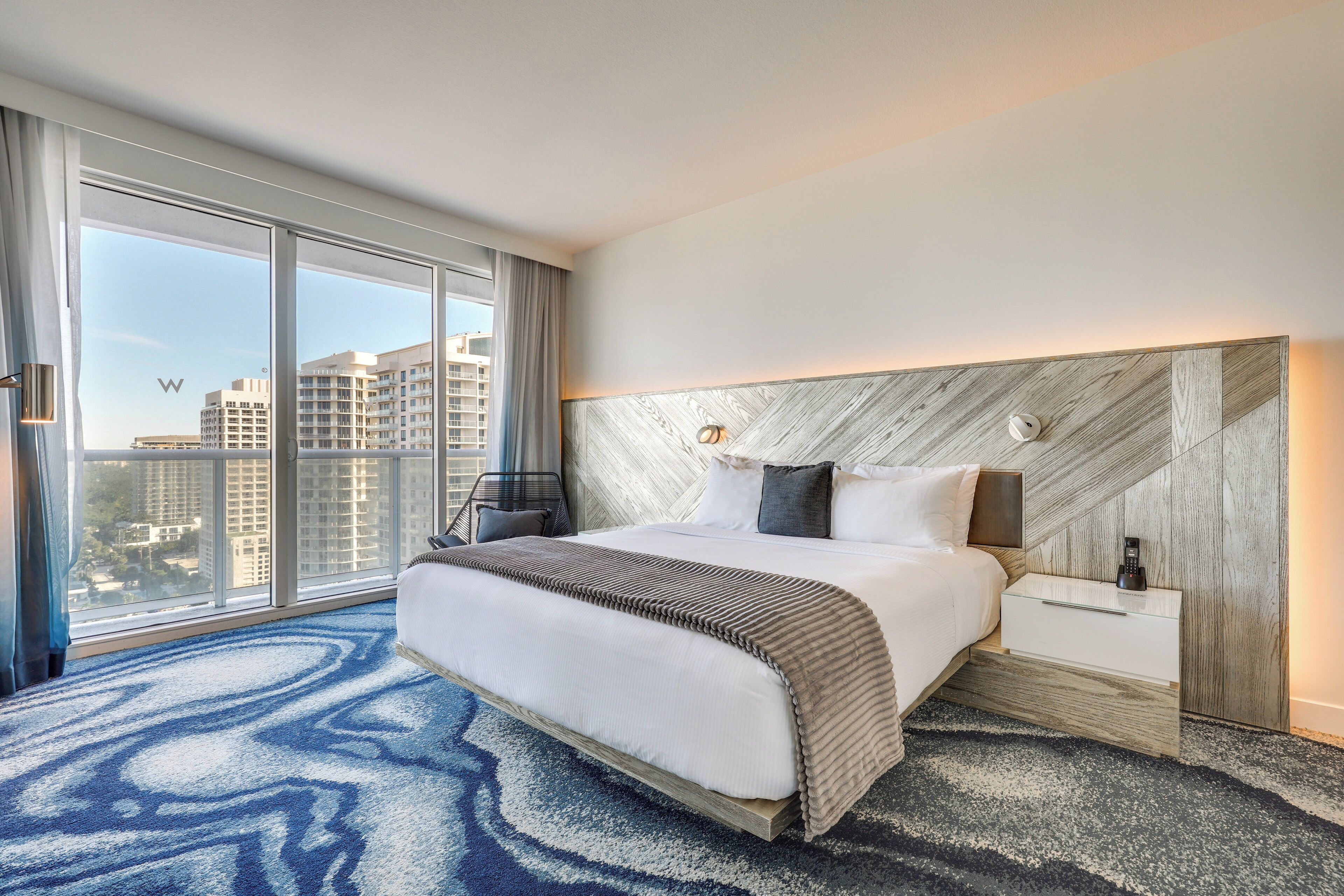 A modern hotel room with a large bed, a nightstand, a lamp, and a view of the city through floor-to-ceiling windows.
