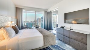 A modern hotel room with a large bed, a flat-screen TV, a city view, and a balcony with a dining area.