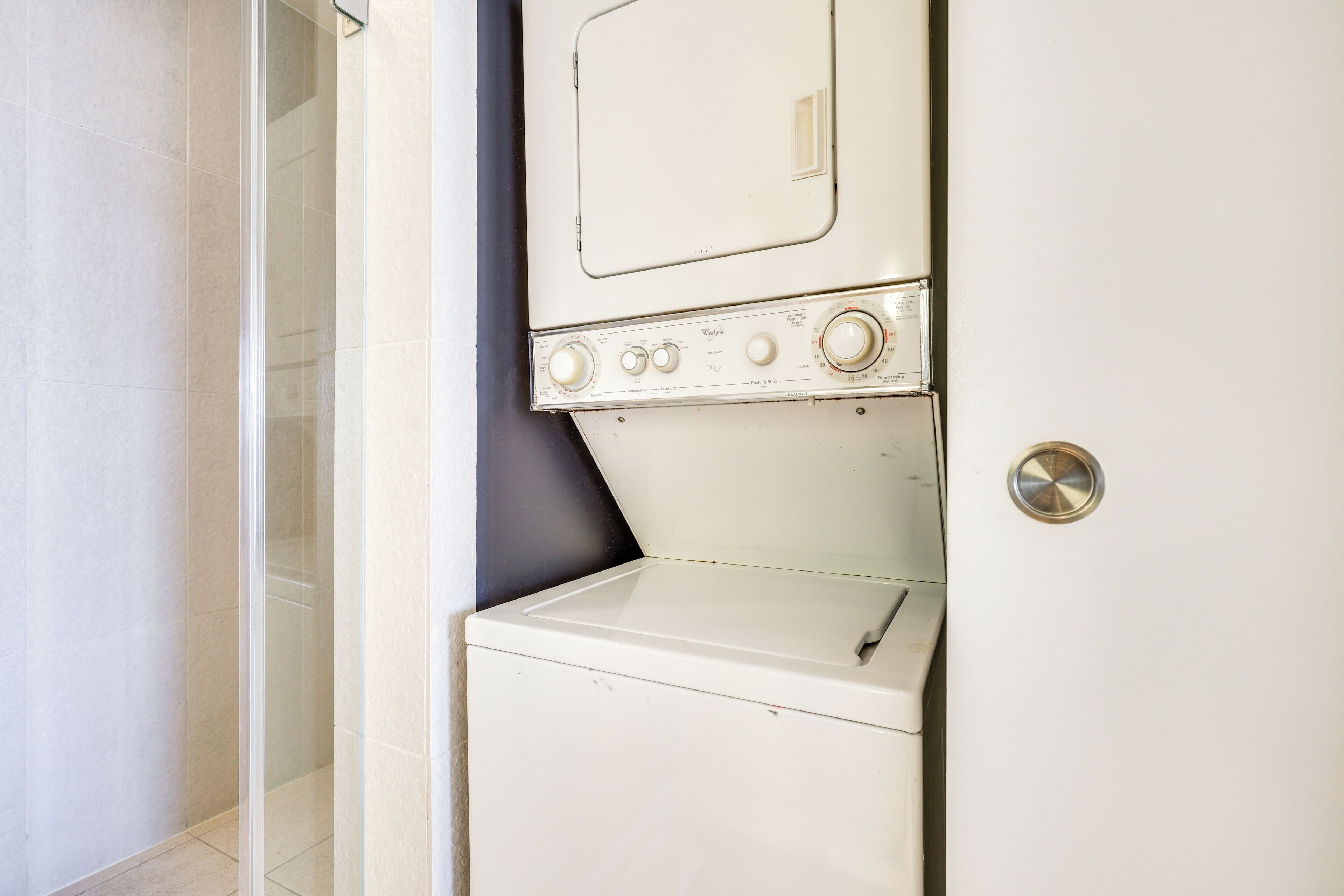 Een kleine, witte wasruimte met een ingebouwde wasmachine en droger, een klein raam en een deur met een ronde hendel.