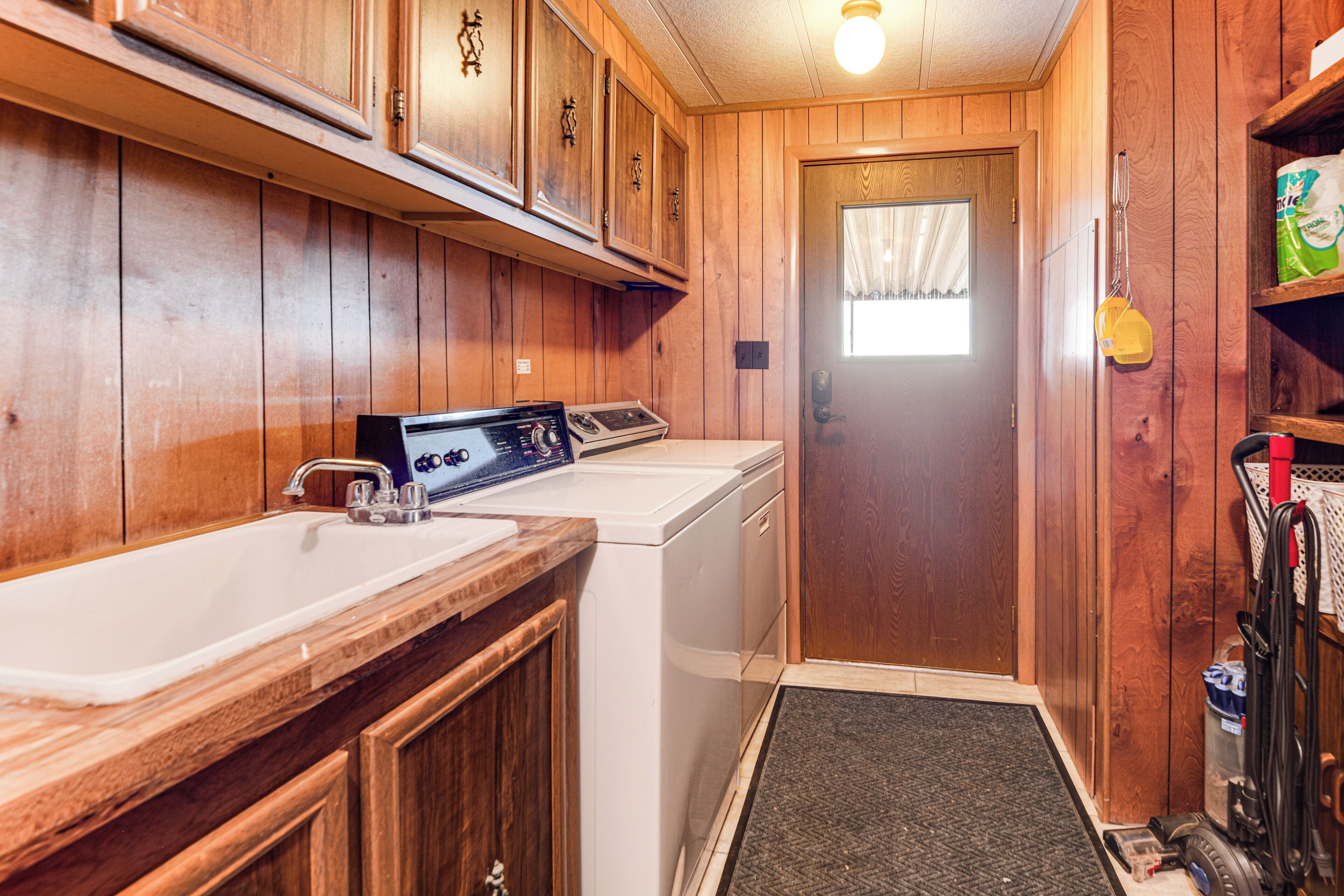 A compact laundry room with wooden walls, a washing machine, a sink, and a door with a window.