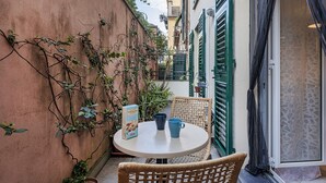 A small outdoor seating area with a round table and wicker chairs, surrounded by a wall with climbing plants and a door with a patterned curtain.