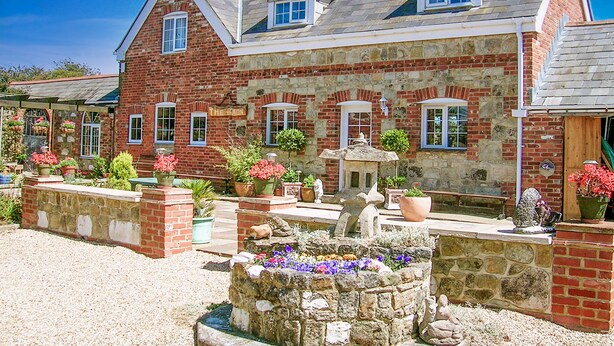 A large, two-story stone building with a sloped roof, multiple windows, and a well-maintained garden with various plants and a decorative stone fountain. - Cosy converted barn in rural location with views of the surrounding countryside. (Newport)