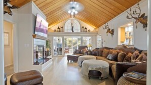 A spacious living room with a high wooden ceiling, a fireplace, and a large television. - Six Suites with Hot Tubs, Private Jacuzzi Baths, Wooded Garden and Lake Access with Boat Slip (Osage Beach)