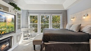 A bedroom with a large bed, a fireplace, a TV, and a view of trees through large windows. - Six Suites with Hot Tubs, Private Jacuzzi Baths, Wooded Garden and Lake Access with Boat Slip (Osage Beach)