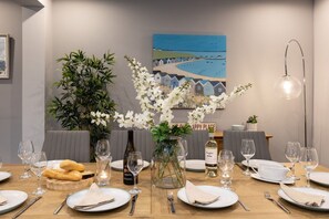 A dining table set with plates, cutlery, glasses, and a bottle of wine, with a vase of flowers and a painting on the wall.
