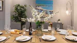 A dining table set with plates, cutlery, glasses, and a bottle of wine, surrounded by chairs and a vase of flowers. - Dwylan : Spacious Family Home Near Rhosneigr Beach | Managed by Anglesey Holiday Lettings (Rhosneigr)