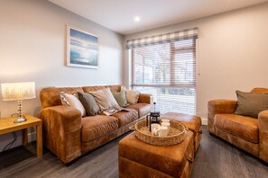A living room with a brown leather sofa, a wooden side table, a wicker coffee table with mugs, and a window with blinds.