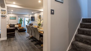 A modern living room with a sofa, chairs, and a dining table. There is a staircase leading to another level. - Dwylan : Spacious Family Home Near Rhosneigr Beach | Managed by Anglesey Holiday Lettings (Rhosneigr)