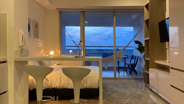 A modern bedroom with a large sliding glass door leading to a balcony, a bed with a white headboard, and a small desk area with a chair. - w Divine Loft w Ocean View in Crespo (Cartagena)