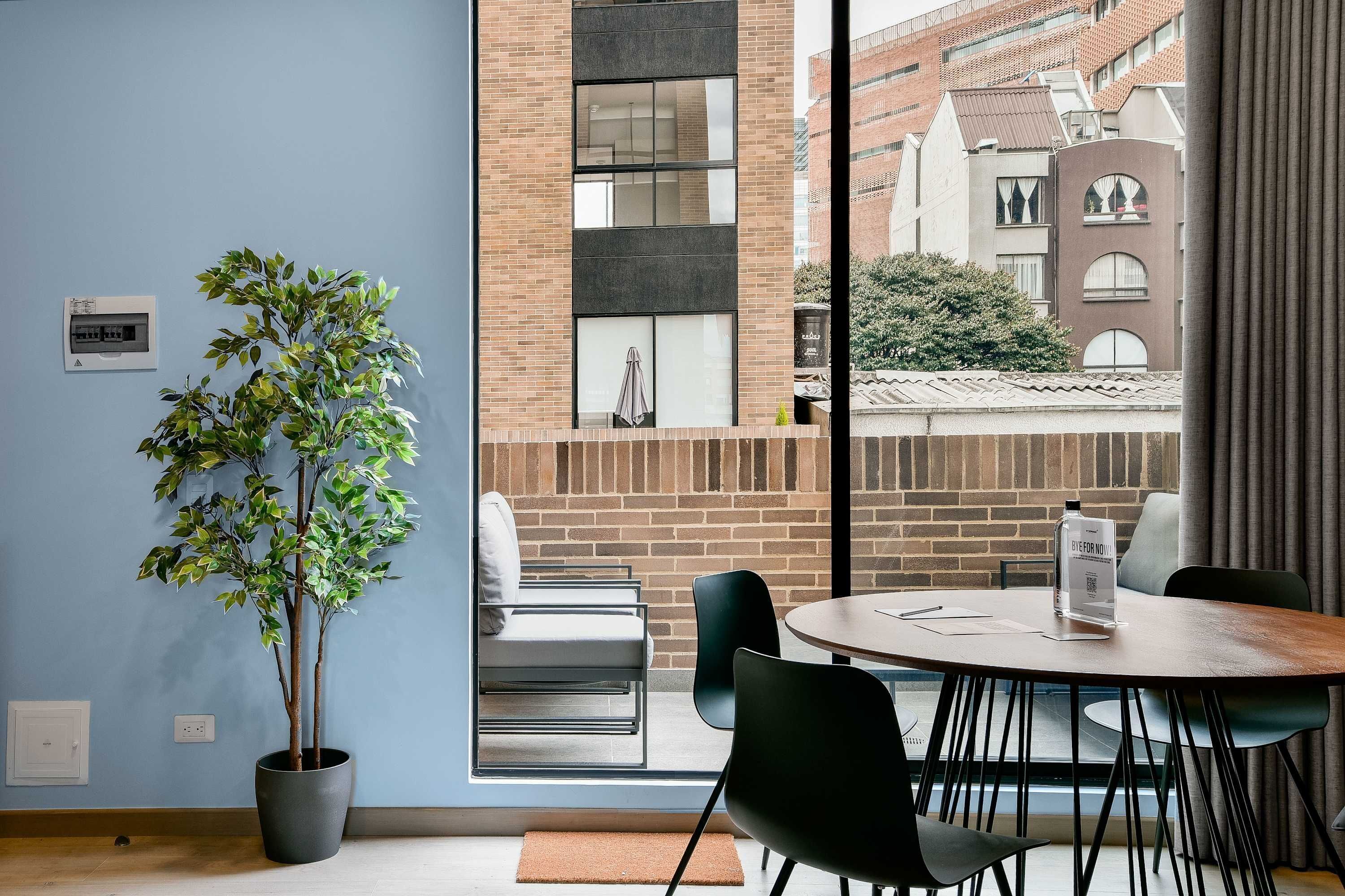 Ein modernes Zimmer mit einem Topfhanf, einem runden Tisch mit Stühlen, einem Sofa und Blick auf ein Ziegelgebäude durch große Fenster.