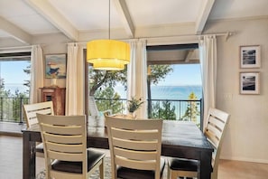 A dining area with a table, chairs, a yellow pendant light, and a view of the sea through large windows. - Cedar Bluff - Gorgeous View! Located in the Tunnel of Trees! Private Beach! (Harbor Springs)