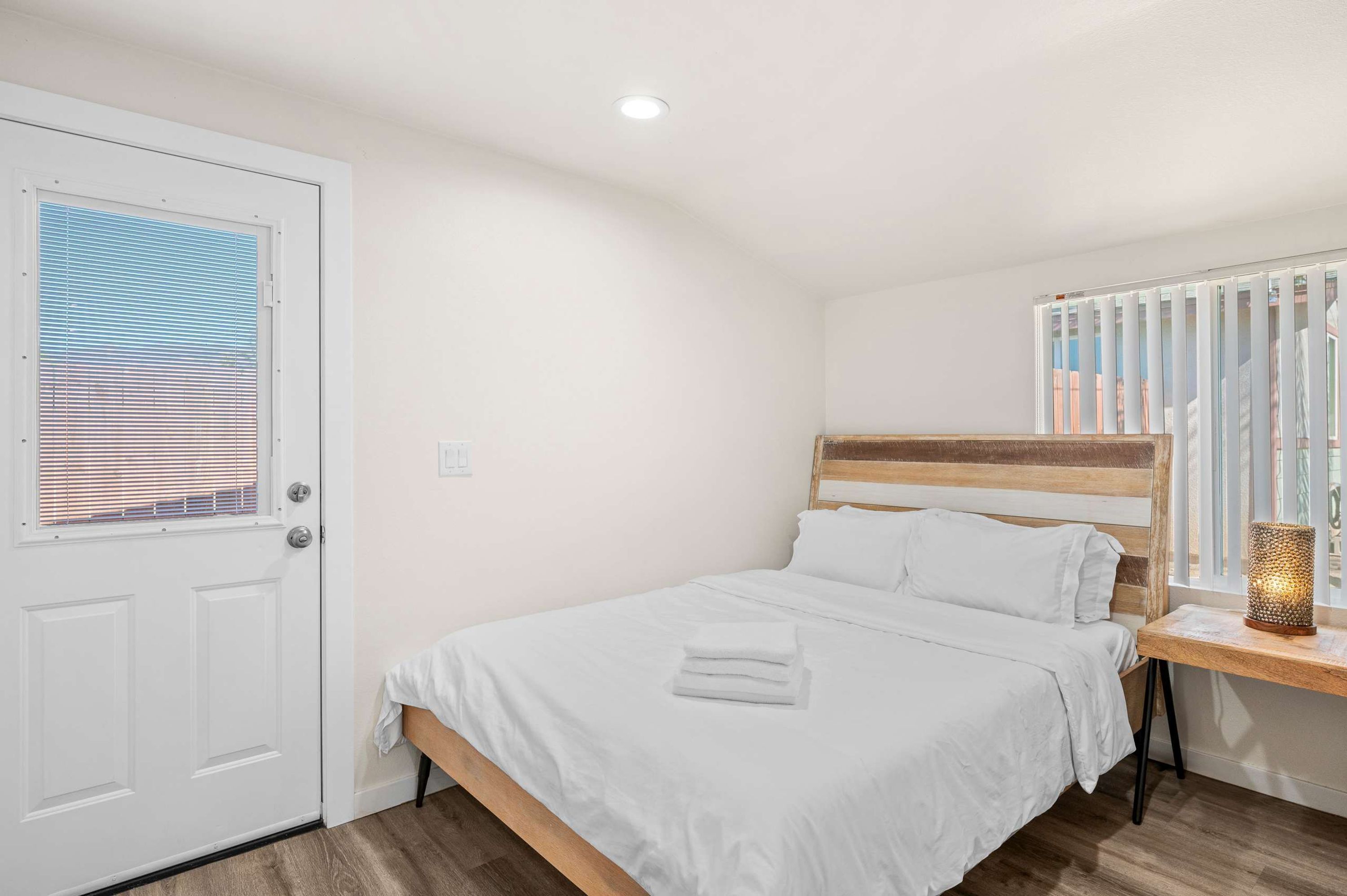 A bedroom with a bed, a wooden headboard, a bedside table with a lamp, and a window with blinds.