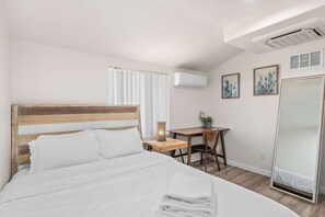 A hotel room with a bed, a desk, a chair, a mirror, and an air conditioning unit. - Charming Escape With Stylish Decor Outdoor Space (Glendora)
