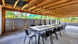 Un espace extérieur abrité, aménagé pour les repas, avec une longue table et des chaises, entouré de rampes en métal et offrant une vue sur la verdure.