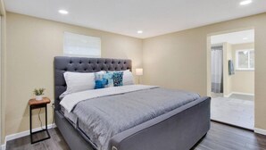 Une chambre à coucher avec une tête de lit capitonnée, une couvre-lit grise et une table de chevet agrémentée d’une plante.