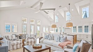 Un salon spacieux avec un plafond haut, de grandes fenêtres et un coin cuisine visible à travers une porte ouverte.