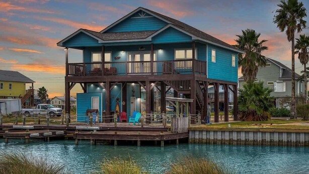 A two-story house with a dock extending into a body of water, surrounded by vegetation. - Twin Palms 2.0 (Rockport)
