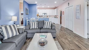 A living room with a gray sofa, striped pillows, a coffee table with a remote, and a kitchen in the background. - Twin Palms 2.0 (Rockport)