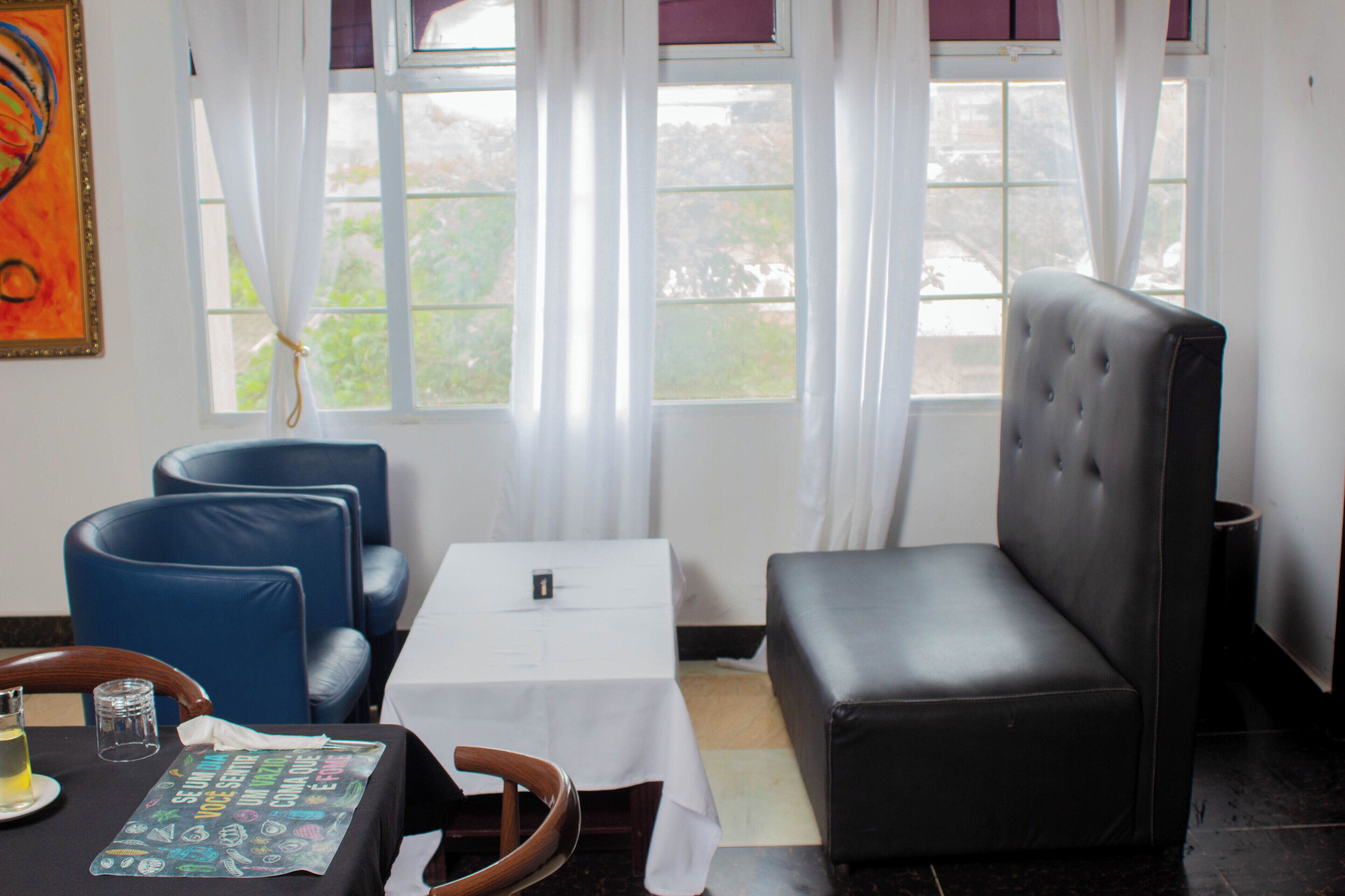 Una habitación con una mesa blanca, sillas azules, un sofá negro y vistas a zonas verdes a través de las ventanas.